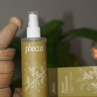 A bottle of phocus Reset Cleanser with a white spray top and olive-green label, displayed on a woven mat next to its matching box and wooden props.