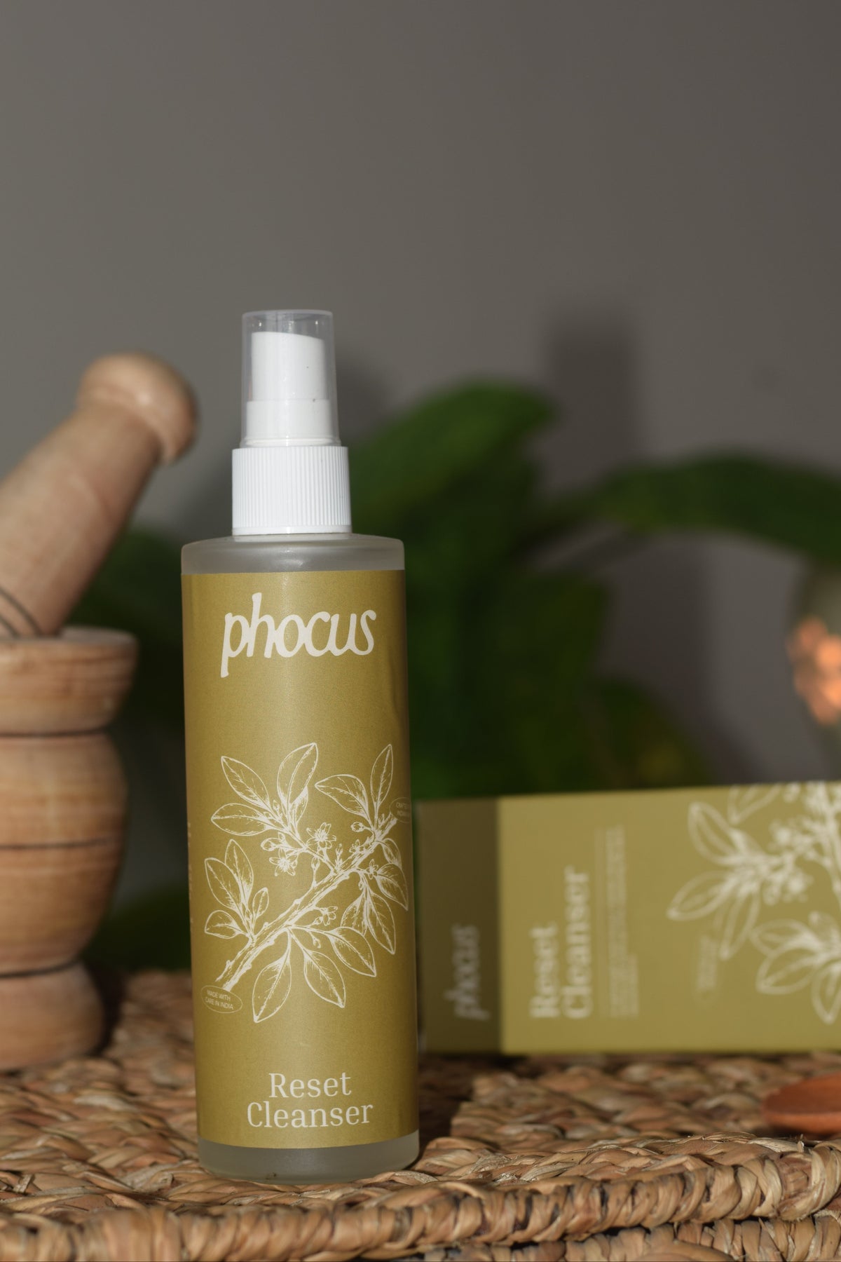 A bottle of phocus Reset Cleanser with a white spray top and olive-green label, displayed on a woven mat next to its matching box and wooden props.