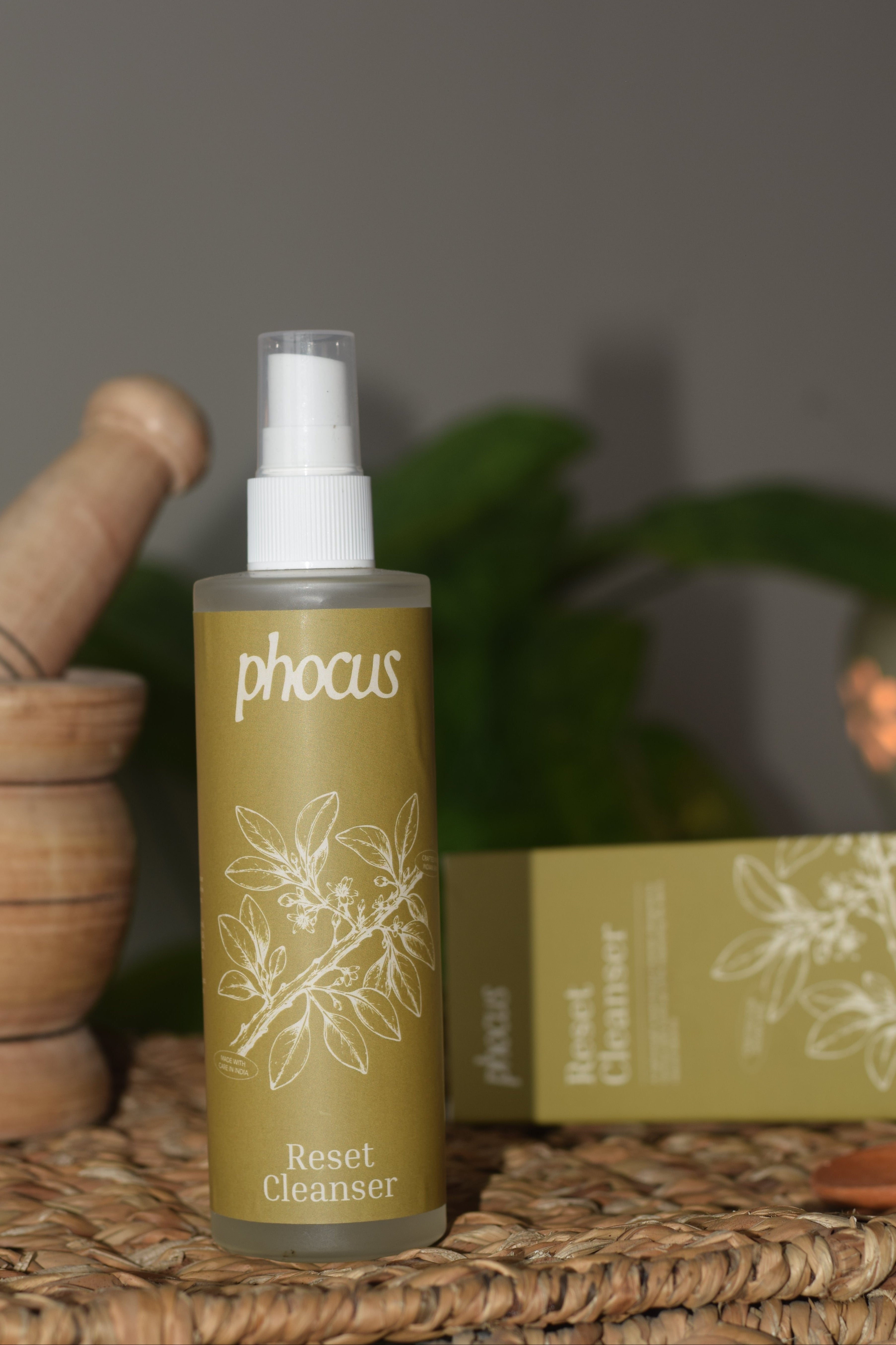 A bottle of phocus Reset Cleanser with a white spray top and olive-green label, displayed on a woven mat next to its matching box and wooden props.
