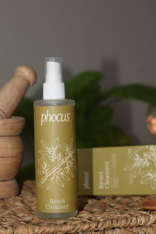 A bottle of phocus Reset Cleanser with a white spray top and olive-green label, displayed on a woven mat next to its matching box and wooden props.